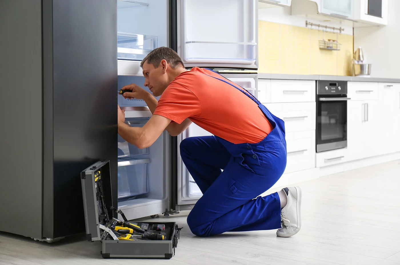 Technician in an orange shirt repairing a refrigerator in a kitchen